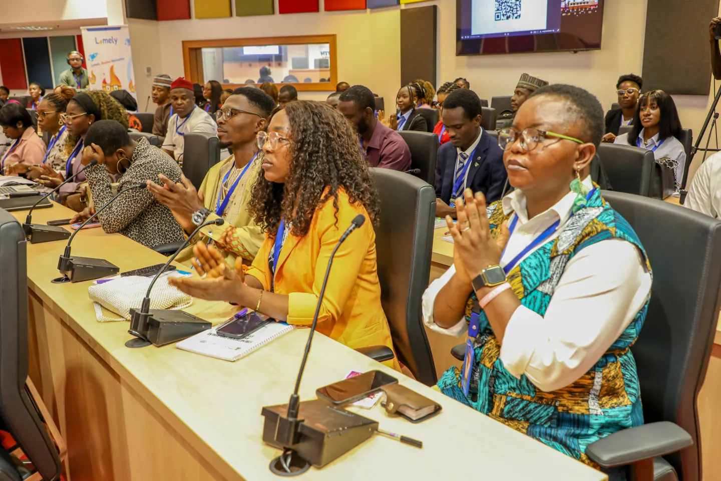 A diverse group of participants seated around conference tables applaud during an event, with microphones, notebooks and mobile phones visible on the desks.