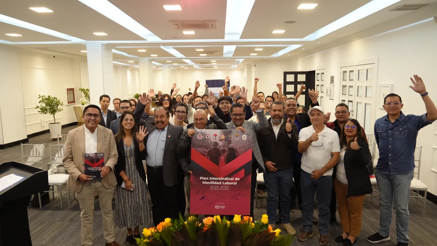 Grupo de personas posan en un salón de eventos y saludan con una mano levantado; mientras las personas en el centro del grupo sostienen un cartel sobre el Plan Intersindical de Movilidad Laboral 2025-2030