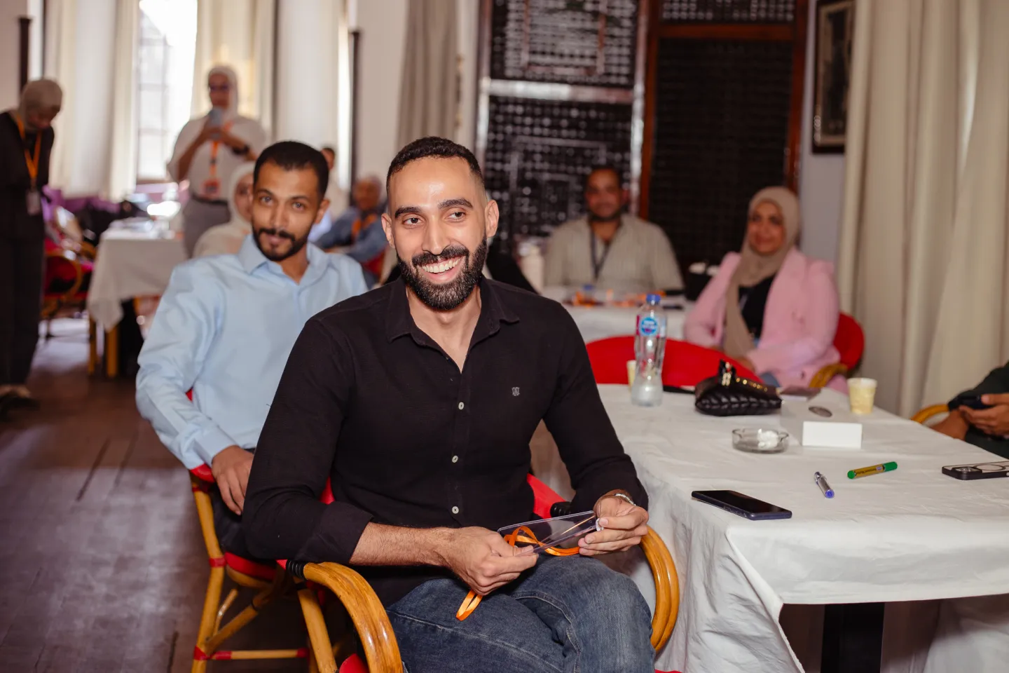 A man seated at a table amongst other seated youth looks into the camera and smiles.