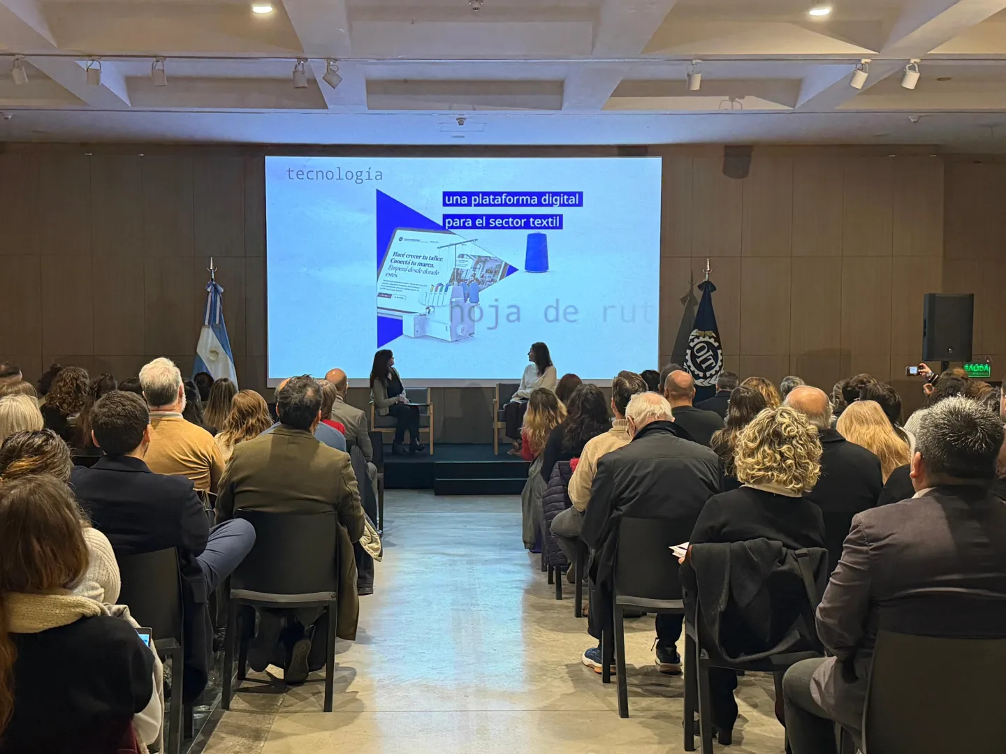 Panel “De la evidencia a la acción” durante el IX Seminario sobre Economía Informal de la OIT en Argentina. Dos expositoras conversan en el escenario frente al público. En la pantalla se presenta una plataforma digital desarrollada para el sector textil.