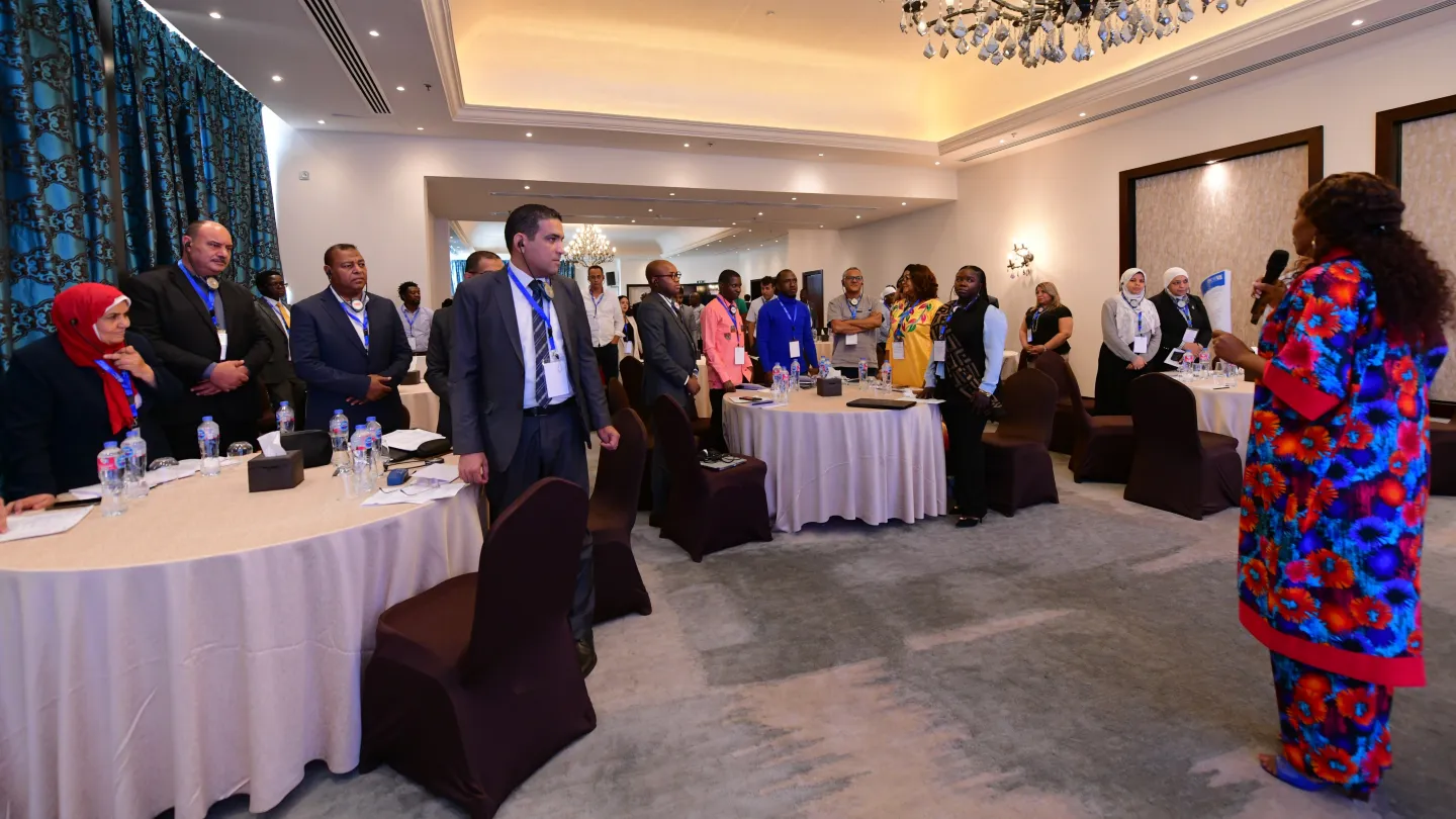 Participants stand around conference tables facing an individual standing in front of them in the middle of the room.