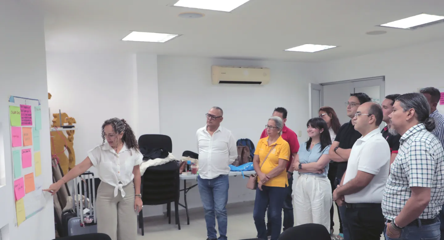 Grupo de personas compuesto por 4 mujeres y 7 hombres observan la cartelera realizada en una pared, que está siendo explicada por una mujer
