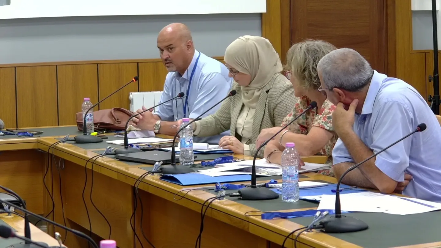 Cinq participants assis autour d’une table de réunion équipée de microphones, de documents et de bouteilles d’eau. Plusieurs prennent des notes tandis qu’un participant, au premier plan, intervient au microphone.
