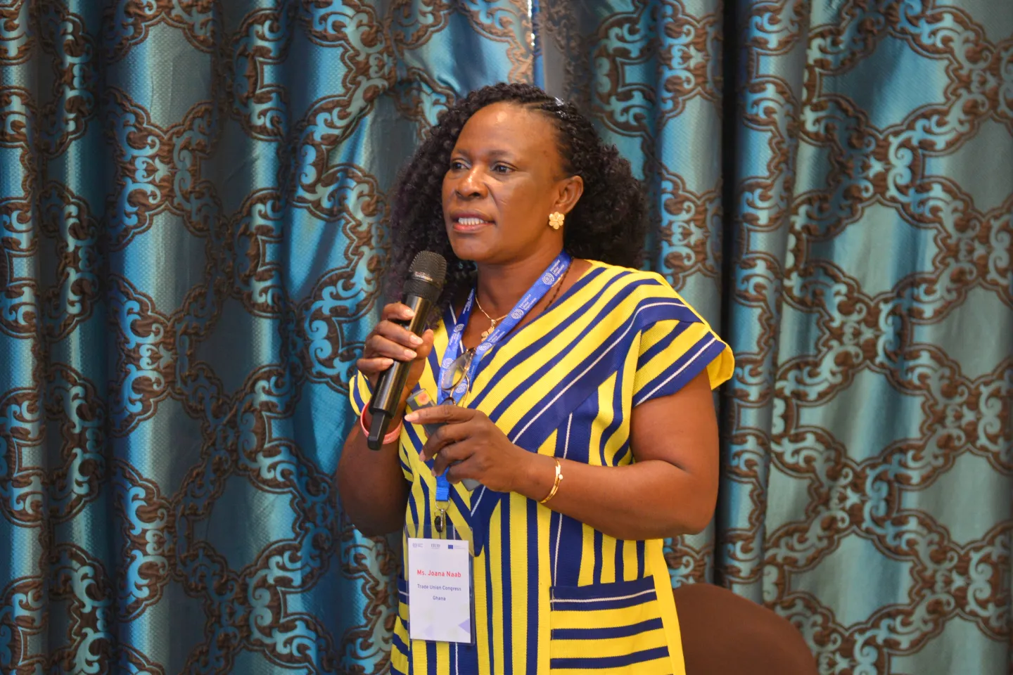 Woman stands in front of patterned curtains and speaks into a handheld microphone