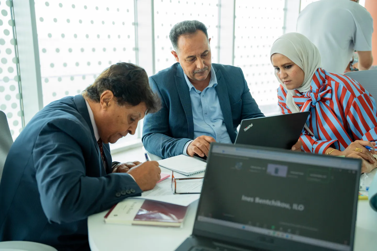 Three participants review documents and information on a laptop during a group discussion