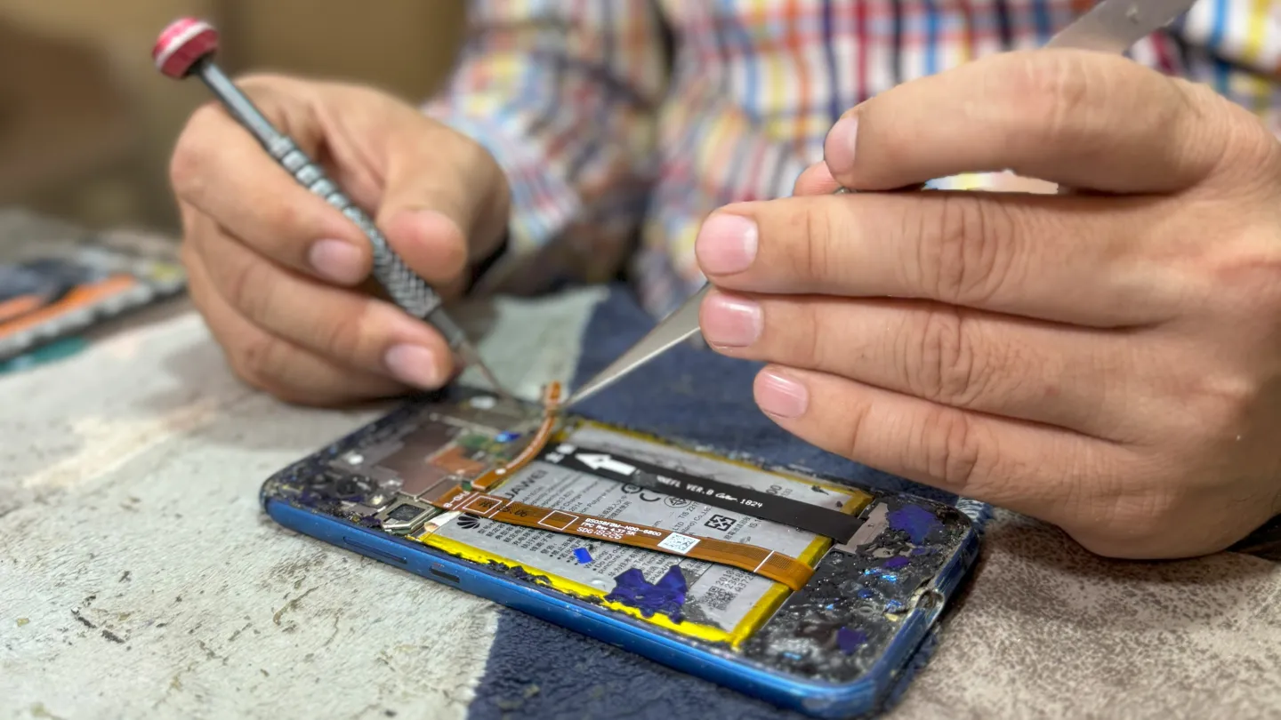 A close-up of Alhassan Ali’s hands holding tools and repairing a mobile phone whose internal mechanism is exposed.