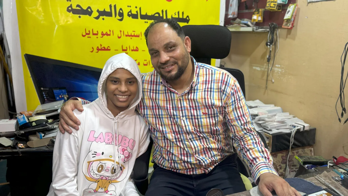Alhassan Ali sits next to his daughter with his arm around her. His daughter wears a white hoodie with the hood up.