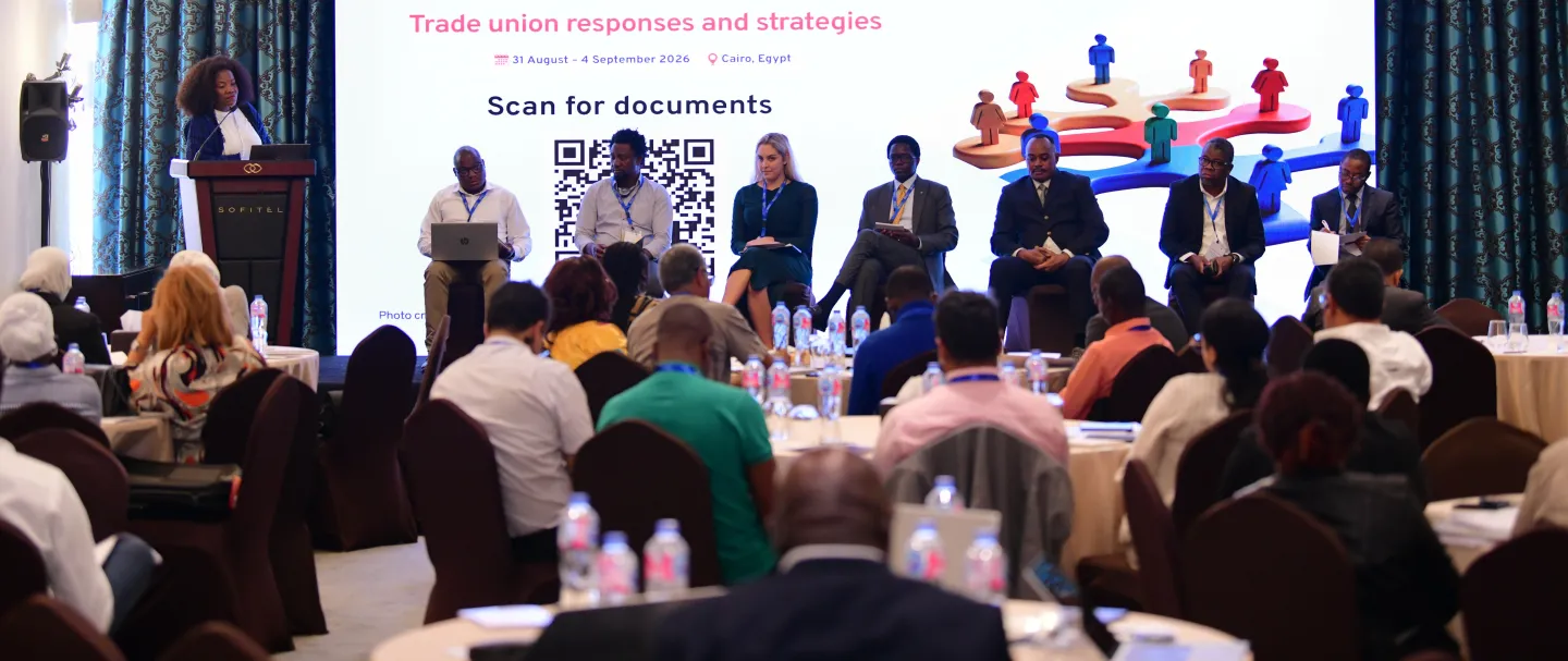 A speaker addresses participants seated in a conference room, with six panellists seated on stage in front of the event screen.