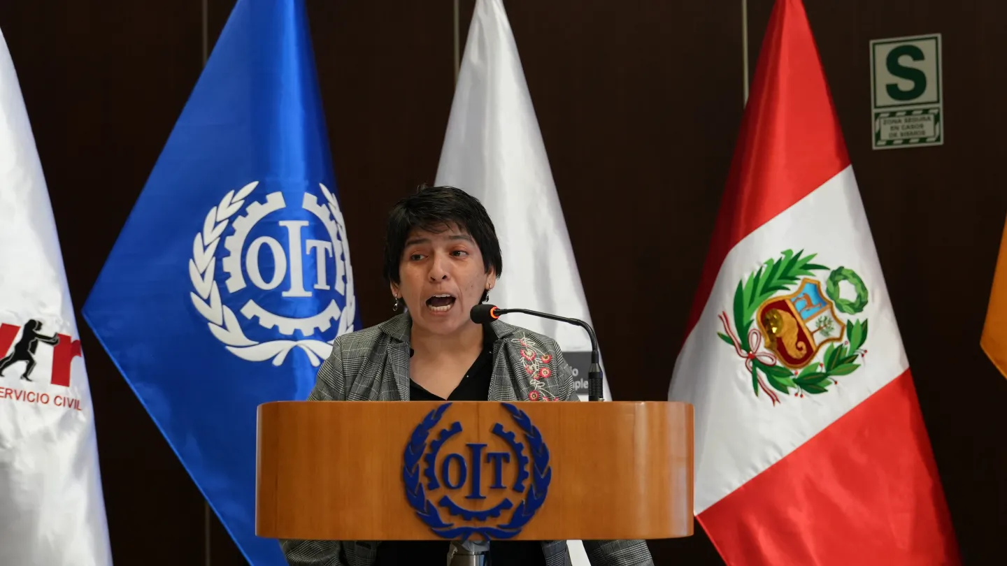 Mujer de cabello corto habla ante un micrófono desde un podio, frente a las banderas de SERVIR, OIT y Perú.