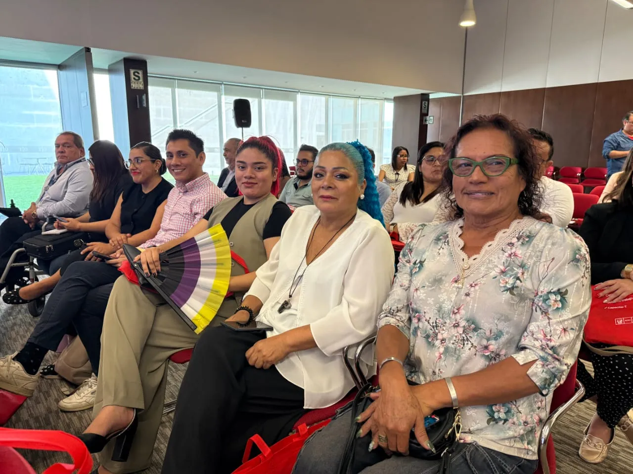Varias personas sentadas en primera fila miran a cámara; una sostiene un abanico con franjas amarillas, blancas, moradas y negras.