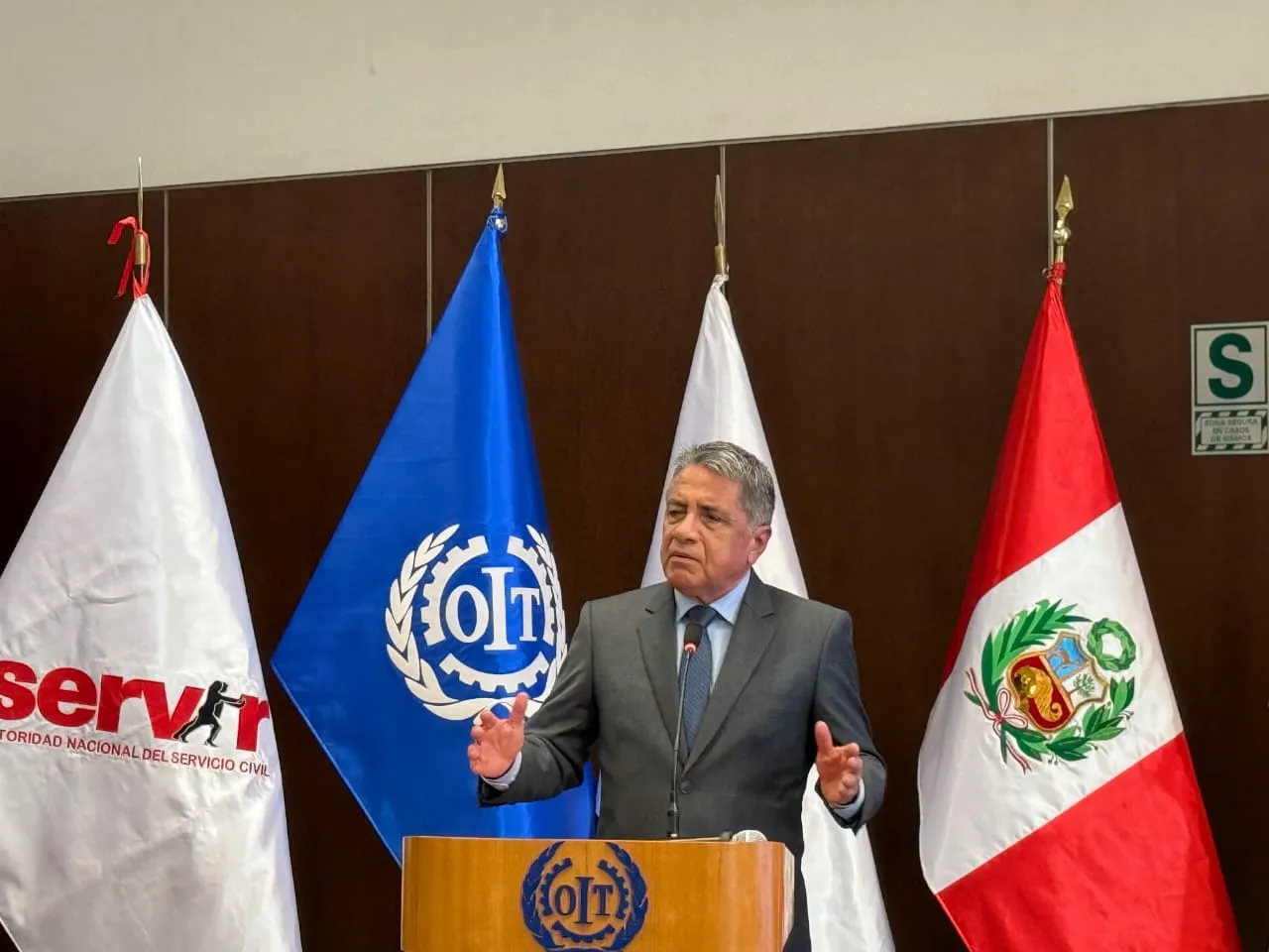 Hombre de traje habla desde un podio, gesticulando con ambas manos, frente a las banderas de SERVIR, OIT y Perú.