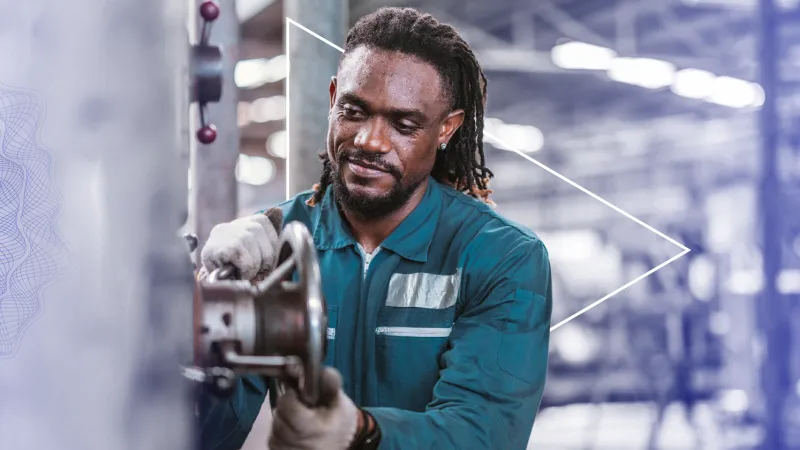 Man wearing a work uniform holds a machine wheel in a factory.