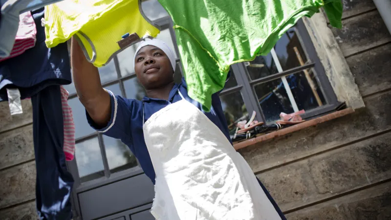 A African woman is hanging colourful clothes on a clothesline outside a stone-walled building with windows.