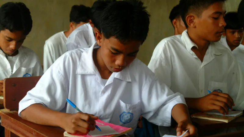 boy in a school uniform writing in a notebook and sitting with his classmates