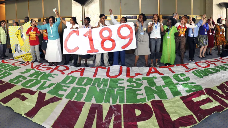 Group of domestic workers, trade union representatives and supporters standing together at the International Labour Conference, holding a large "C189" sign and a banner calling for ratification and implementation of the Domestic Workers Convention. Participants raise joined hands in celebration following the adoption of Convention No. 189 on decent work for domestic workers.